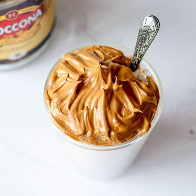 A glass of whipped Dalgona coffee with a layer of frothy coffee cream on top of milk, served with a spoon. A jar of instant coffee is partially visible in the background.