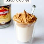 A clear glass filled with milk topped with fluffy whipped coffee, with a spoon sticking out, and a blurred jar of instant coffee in the background.
