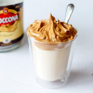 A clear glass filled with milk topped with fluffy whipped coffee, with a spoon sticking out, and a blurred jar of instant coffee in the background.