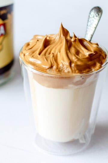 A clear glass filled with milk topped with fluffy whipped coffee, with a spoon sticking out, and a blurred jar of instant coffee in the background.