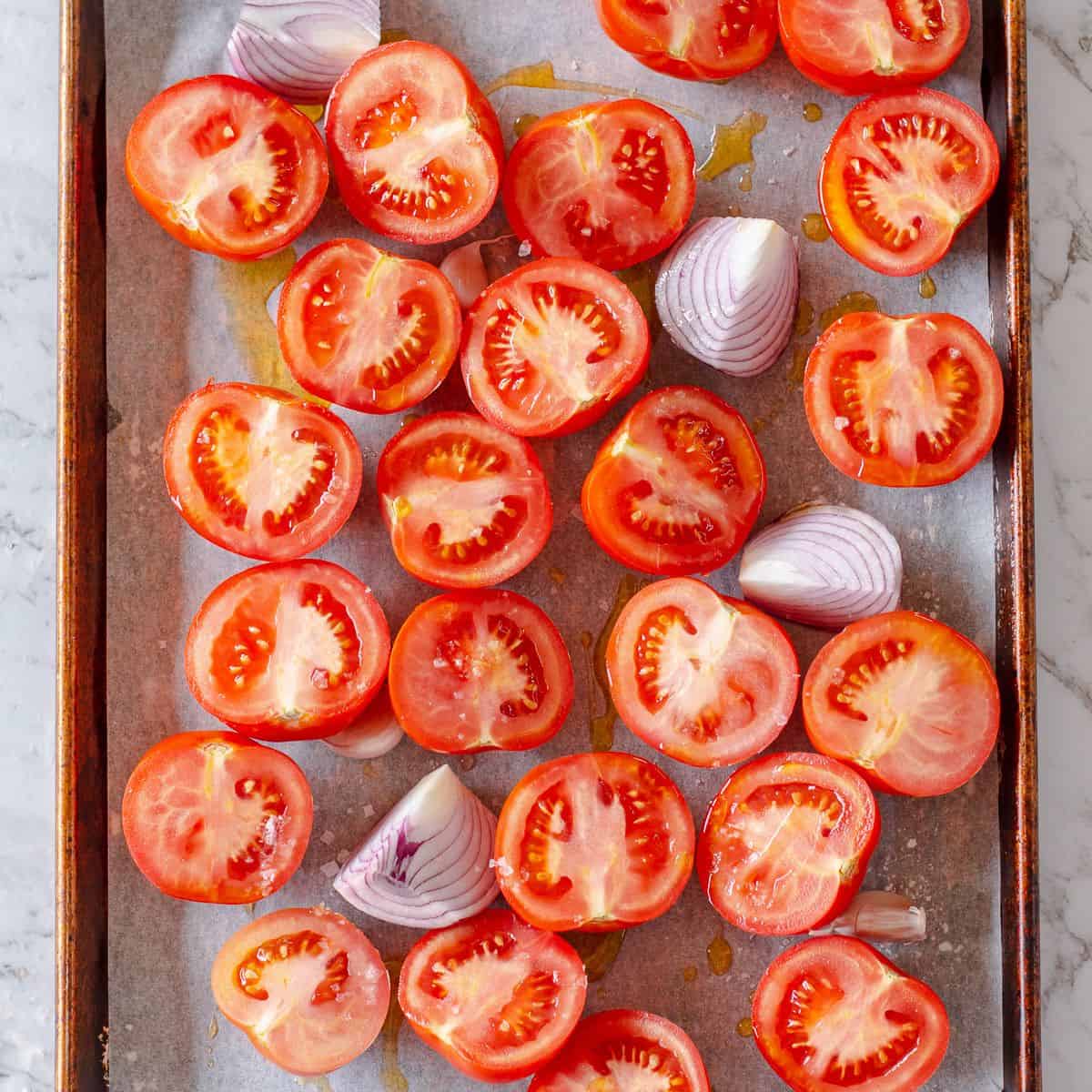 Halved tomatoes and quartered red onions arranged on a baking tray lined with baking parchment, drizzled with olive oil, ready for roasting.