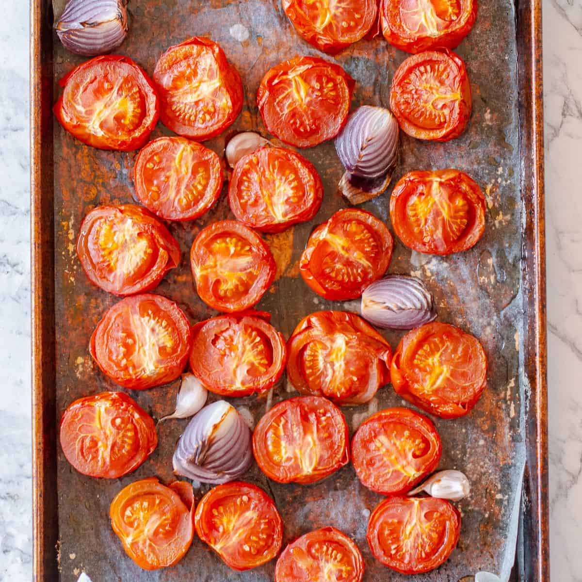 A baking tray with halved roasted tomatoes, sections of red onion, and garlic cloves on greaseproof paper, set on a marble worktop.