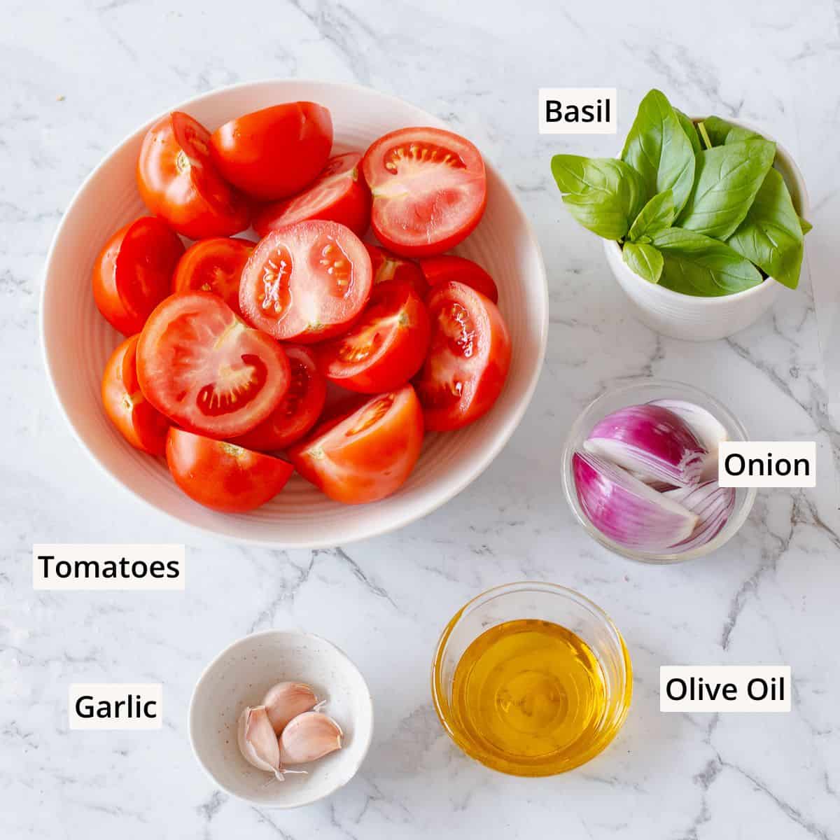 A marble surface with bowls containing halved tomatoes, fresh basil leaves, sliced red onion, garlic cloves, and a small bowl of olive oil. Each ingredient is labelled with text.