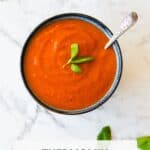 Tomato and basil soup in a blue bowl on a white marble bench top.