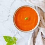 A bowl of smooth tomato soup topped with a basil leaf sits on a marble surface next to a kitchen tea towel, with fresh basil leaves placed nearby.