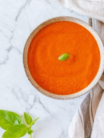 A bowl of smooth tomato soup topped with a basil leaf sits on a marble surface next to a kitchen tea towel, with fresh basil leaves placed nearby.