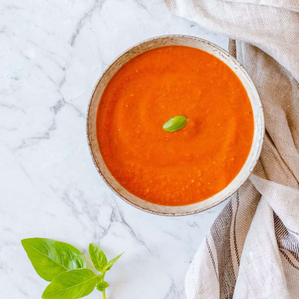A bowl of smooth tomato soup topped with a basil leaf sits on a marble surface next to a kitchen tea towel, with fresh basil leaves placed nearby.