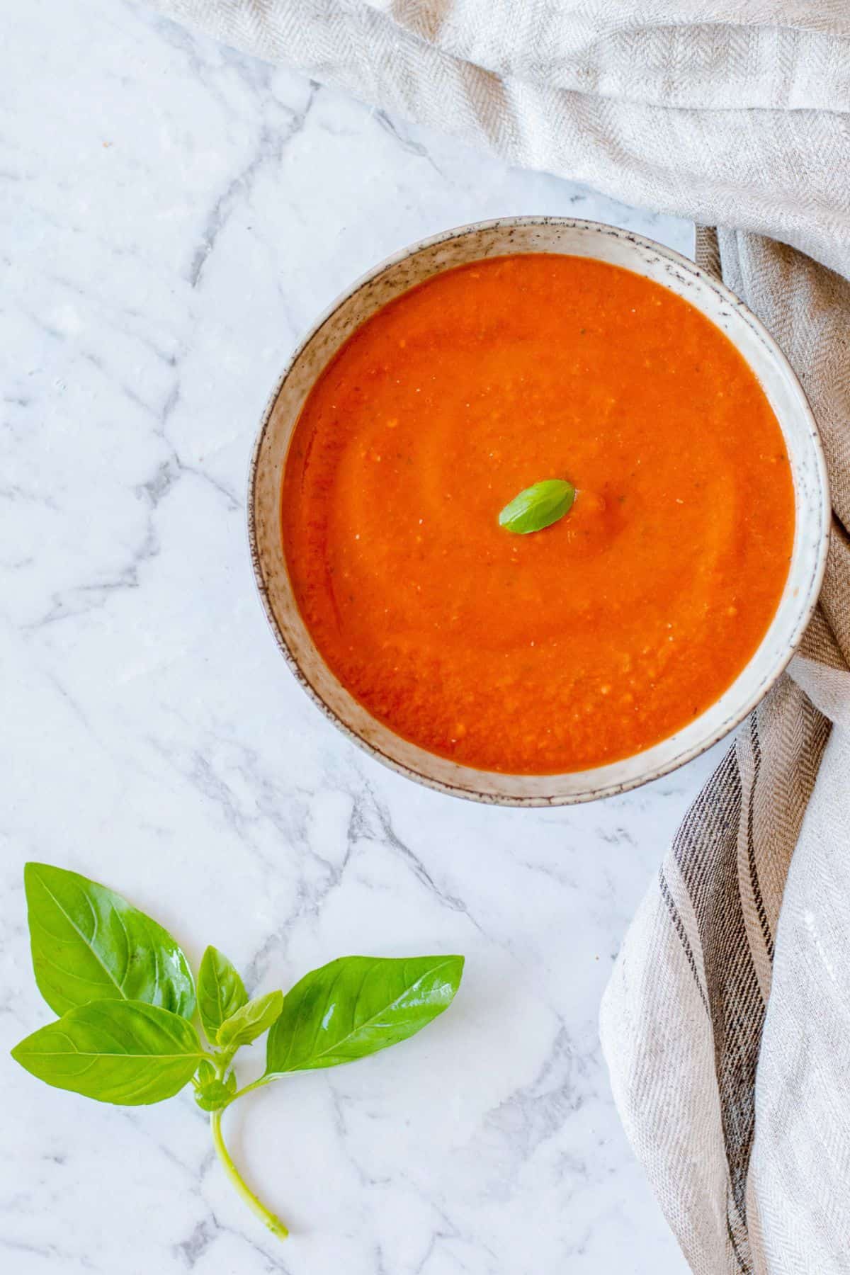 A bowl of smooth tomato soup topped with a basil leaf sits on a marble surface next to a kitchen tea towel, with fresh basil leaves placed nearby.