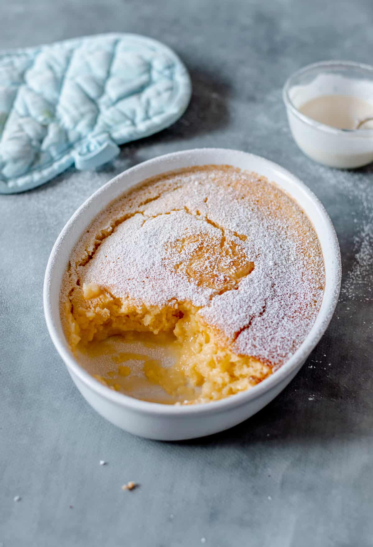 Oval shaped bowl of Lemon Delicious Pudding half eaten on blue background with oven glove and cream