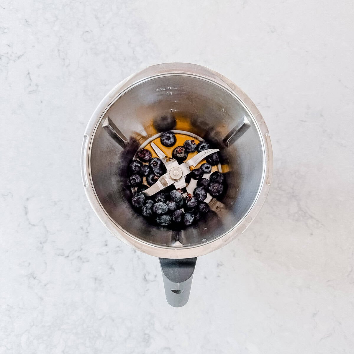 A top-down view of a thermomix bowl containing a small amount of blueberries and a bit of liquid, placed on a light-coloured, marble-like surface.