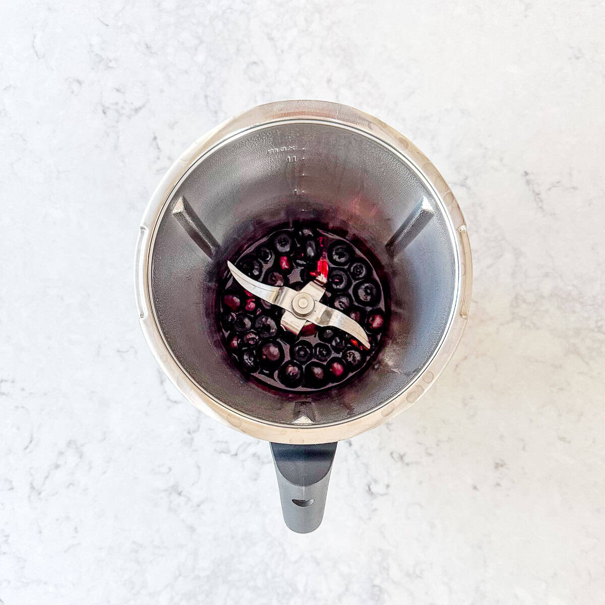 A top-down view of a thermomix bowl with cooked blueberries inside, set on a light-coloured marble countertop.
