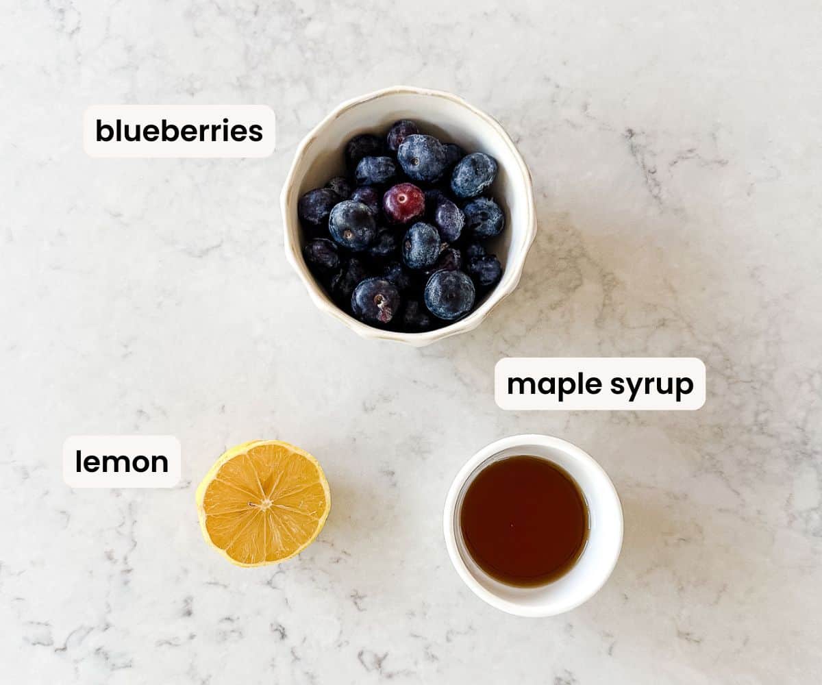A bowl of blueberries, a small cup of maple syrup, and a halved lemon are arranged on a light marble surface. Each item is labelled with text.