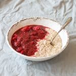 Apple and Raspberry Compote with porridge in a white bowl, with spoon, sitting on a grey tablecloth.