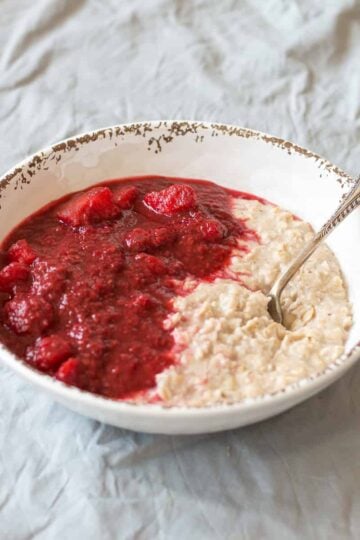 Apple and Raspberry Compote with porridge in a white bowl, with spoon, sitting on a grey tablecloth.