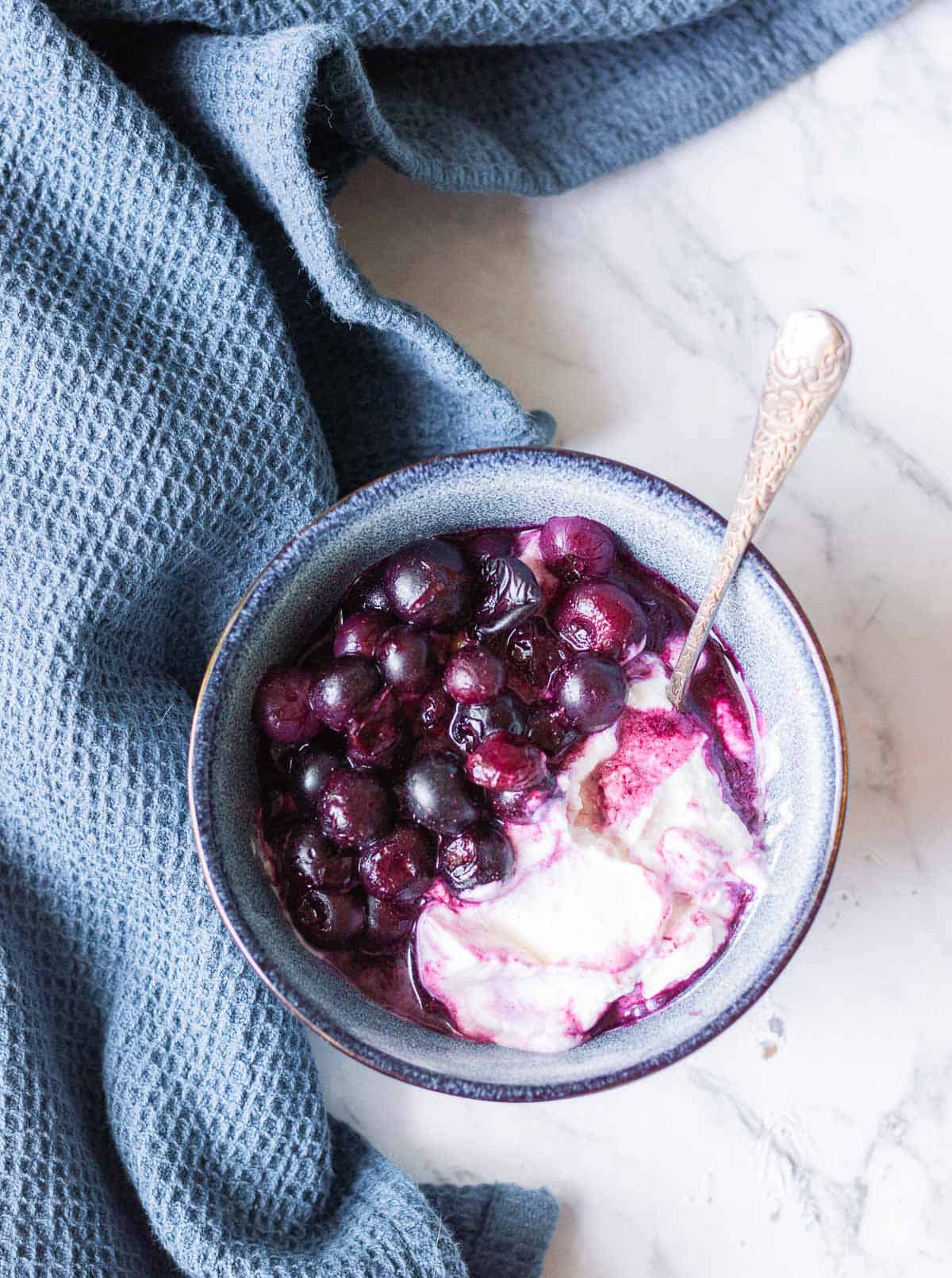 A bowl of yoghurt topped with cooked blueberries sits on a marble surface next to a textured blue cloth. A spoon is placed in the bowl.