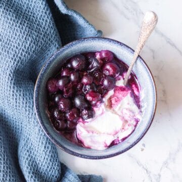 A bowl of yoghurt topped with cooked blueberries sits on a marble surface next to a textured blue cloth. A spoon is placed in the bowl.