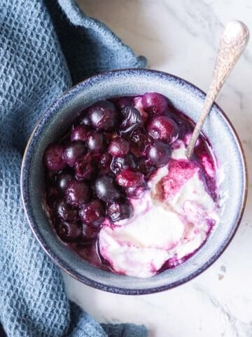 A bowl of yoghurt topped with cooked blueberries sits on a marble surface next to a textured blue cloth. A spoon is placed in the bowl.