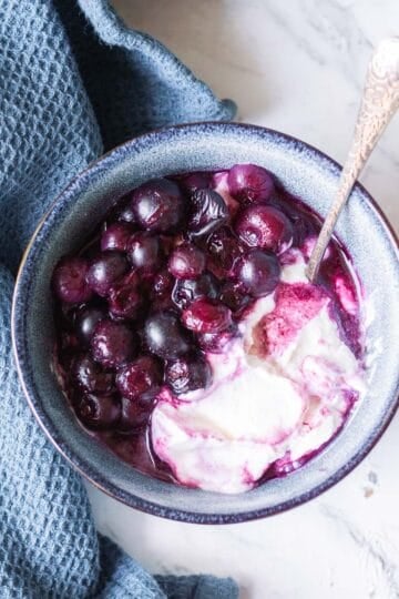 A bowl of yoghurt topped with cooked blueberries sits on a marble surface next to a textured blue cloth. A spoon is placed in the bowl.