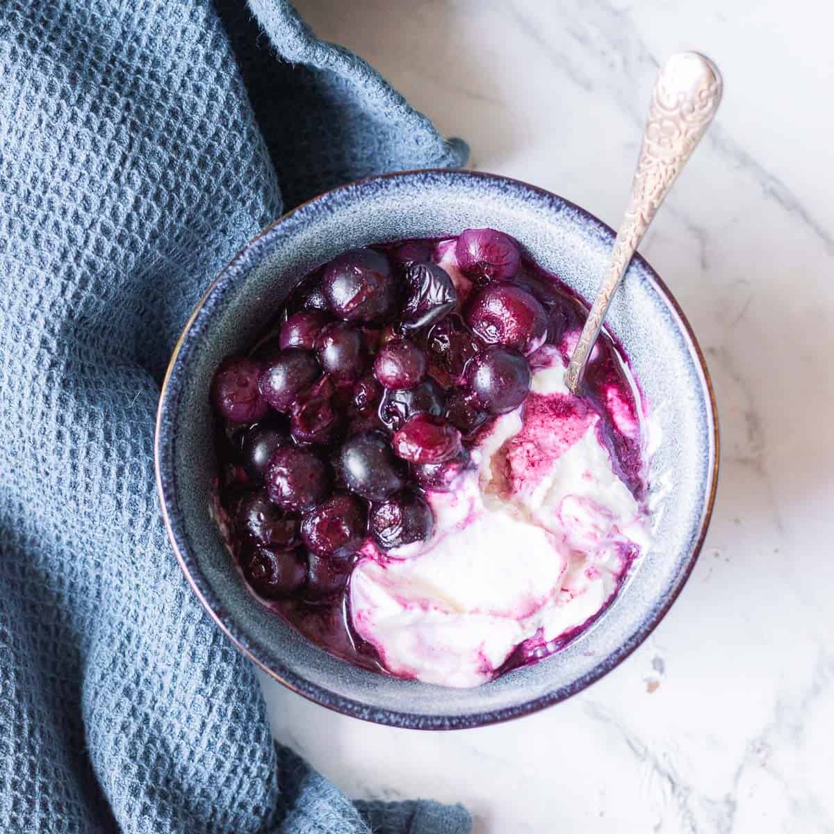 A bowl of yoghurt topped with cooked blueberries sits on a marble surface next to a textured blue cloth. A spoon is placed in the bowl.