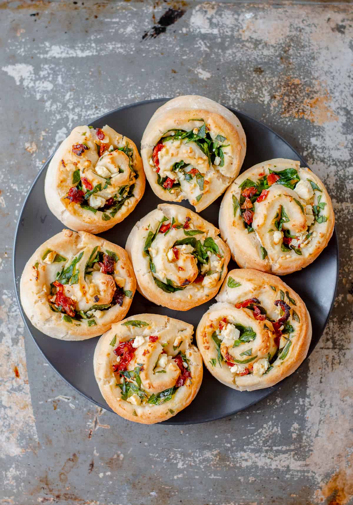 Seven spinach, feta and sundried tomato scrolls on a black plate on grey background.