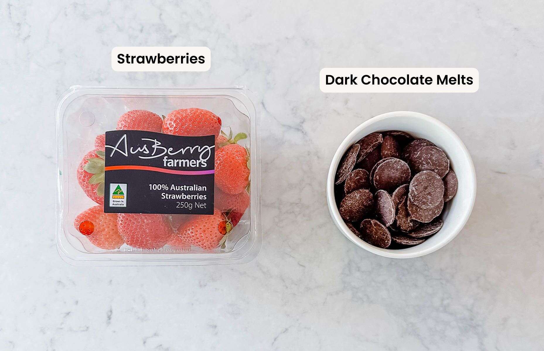 A clear plastic container of fresh strawberries sits on the left, labelled AusBerry farmers, next to a white bowl filled with dark chocolate melts on a light marble surface. Labels indicate Strawberries and Dark Chocolate Melts.