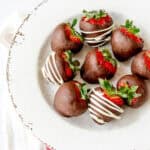A white plate with seven chocolate-covered strawberries, some drizzled with white chocolate, sits on a white cloth with a red stripe.