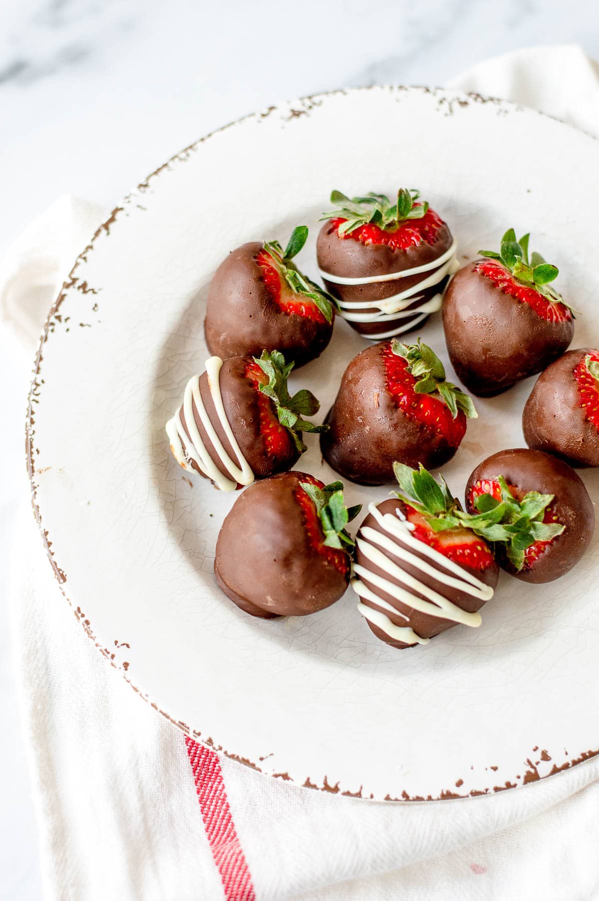 A white plate with seven chocolate-covered strawberries, some drizzled with white chocolate, sits on a white cloth with a red stripe.