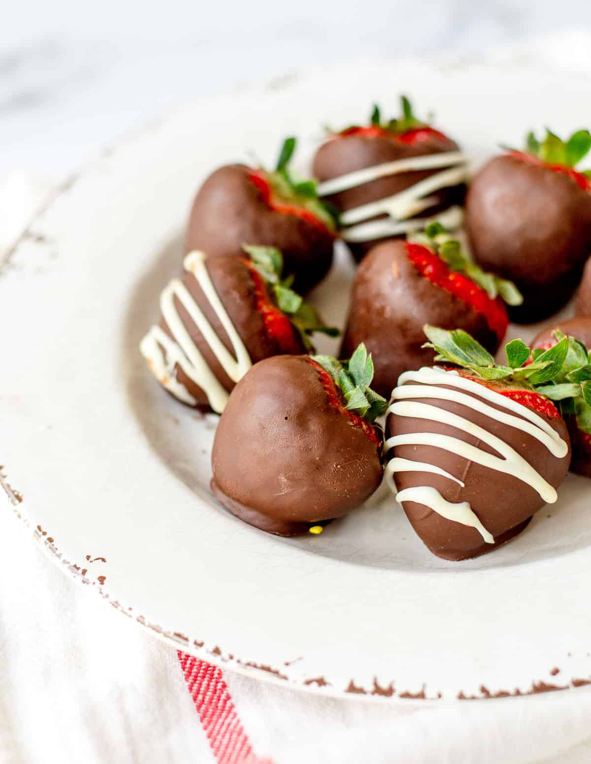A white plate with chocolate-covered strawberries, some drizzled with white chocolate, arranged on a light cloth with a red stripe.