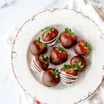 A white plate with a rustic, chipped edge holds seven strawberries dipped in chocolate. Some are drizzled with white chocolate, and all have green tops intact. The plate rests on a white cloth.