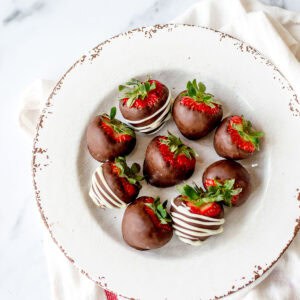 A white plate with a rustic, chipped edge holds seven strawberries dipped in chocolate. Some are drizzled with white chocolate, and all have green tops intact. The plate rests on a white cloth.