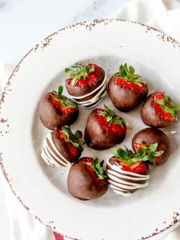 A white plate with a rustic, chipped edge holds seven strawberries dipped in chocolate. Some are drizzled with white chocolate, and all have green tops intact. The plate rests on a white cloth.