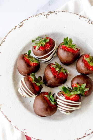 A white plate with a rustic, chipped edge holds seven strawberries dipped in chocolate. Some are drizzled with white chocolate, and all have green tops intact. The plate rests on a white cloth.