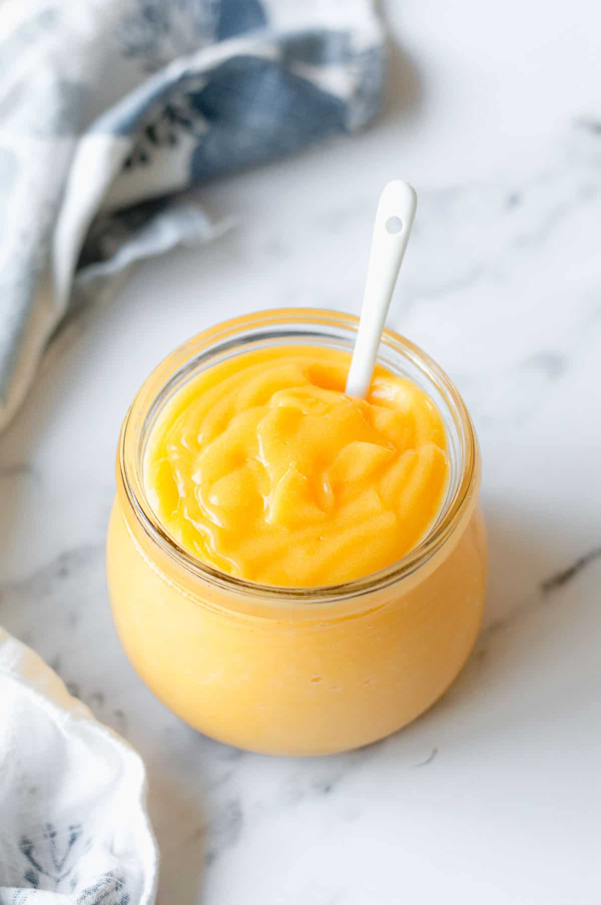 Lemon curd in a glass jar with white spoon and bluey white tea towel.