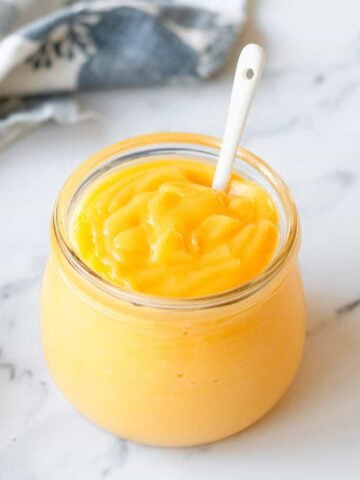 Lemon Curd in a glass jar with white spoon and blue/white tea towel.