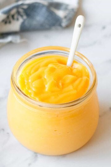 Lemon Curd in a glass jar with white spoon and blue/white tea towel.