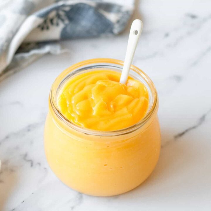 Lemon Curd in a glass jar with white spoon and blue/white tea towel.