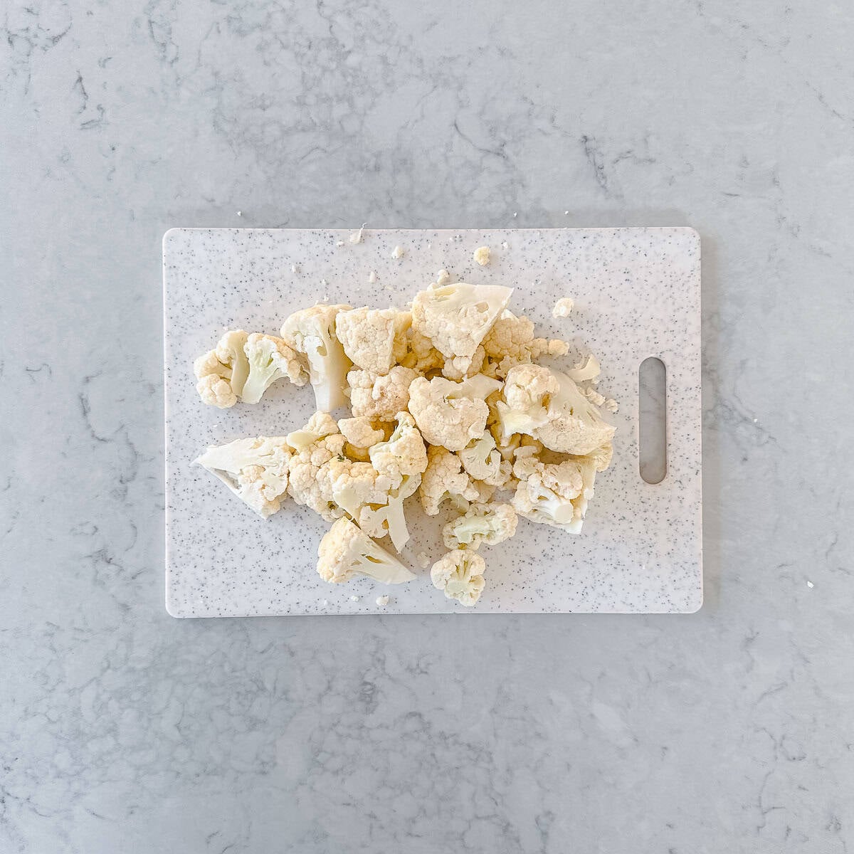 Chopped cauliflower florets are spread across a white speckled chopping board on a light grey marble surface.