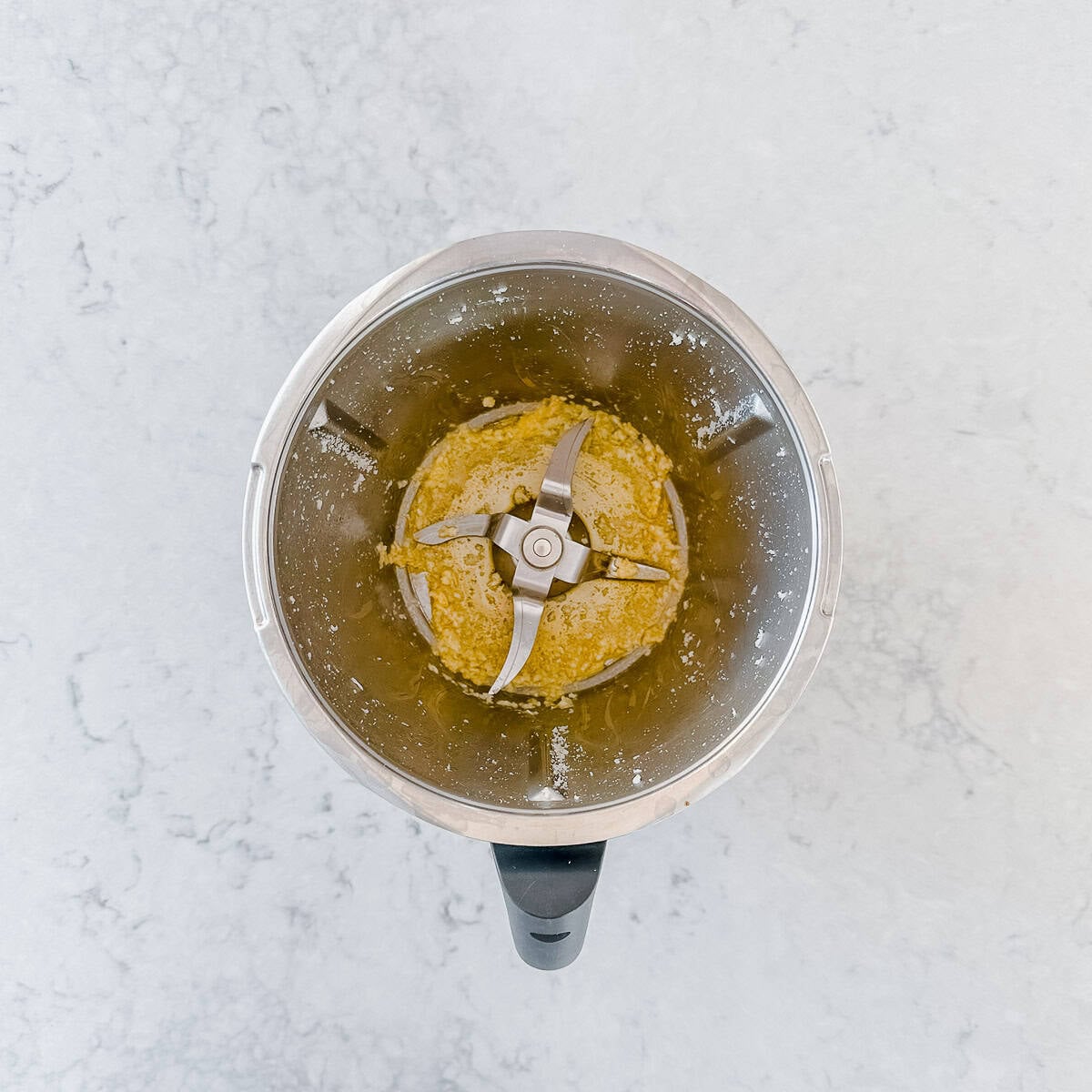 Top-down view of a Thermomix with sautéd garlic inside, sitting on a light marbled worktop. The metal blades are visible, surrounded by the blended ingredients.
