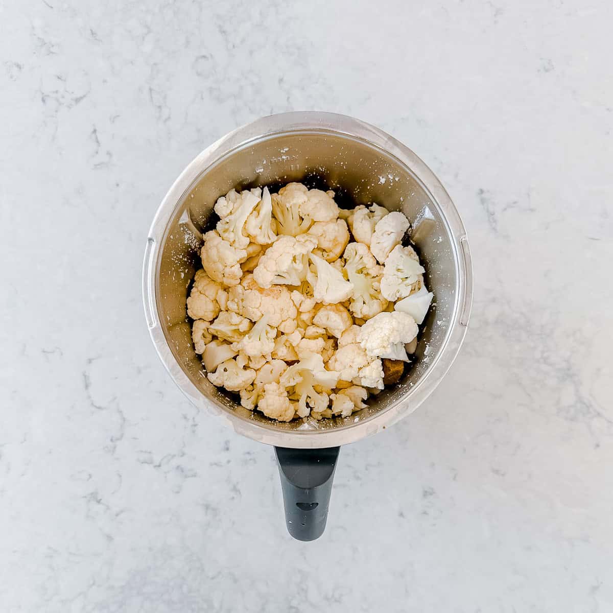 A top-down view of a Thermomix bowl filled with raw cauliflower florets on a light, marble-like worktop.