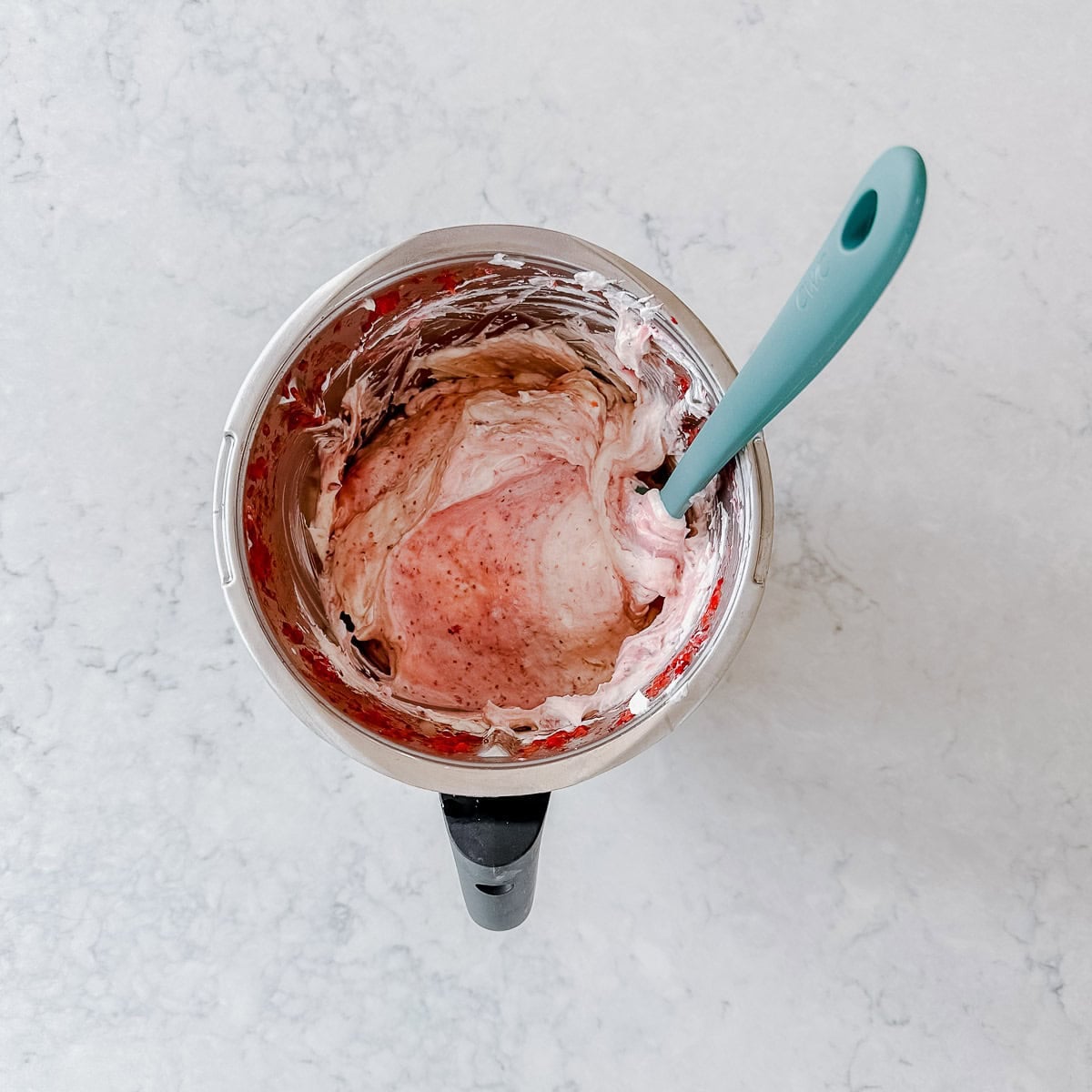 A mixing bowl with pink and white creamy mixture being stirred by a teal spatula, placed on a light marble surface.