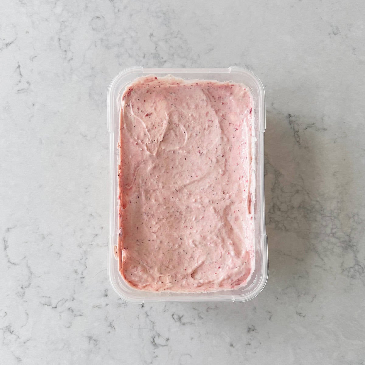 A rectangular plastic container filled with light pink, creamy spread, sitting on a white and gray marbled surface. The spread appears smooth with small red specks throughout.