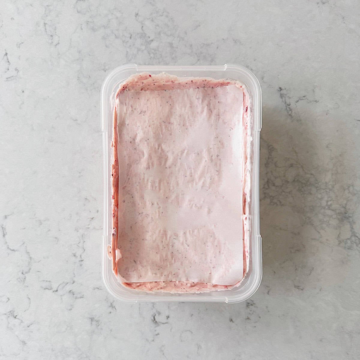 A rectangular plastic container filled with a smooth, pink, creamy strawberry ice cream placed on a light grey marbled surface.