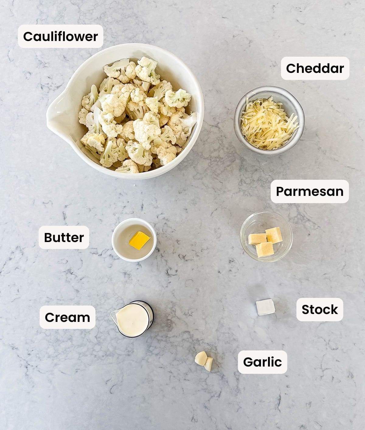 Ingredients for a cauliflower cheese soup laid out on a counter: bowl of cauliflower florets, grated cheddar, grated parmesan, butter, cream, stock cube, and two garlic cloves, each labelled.