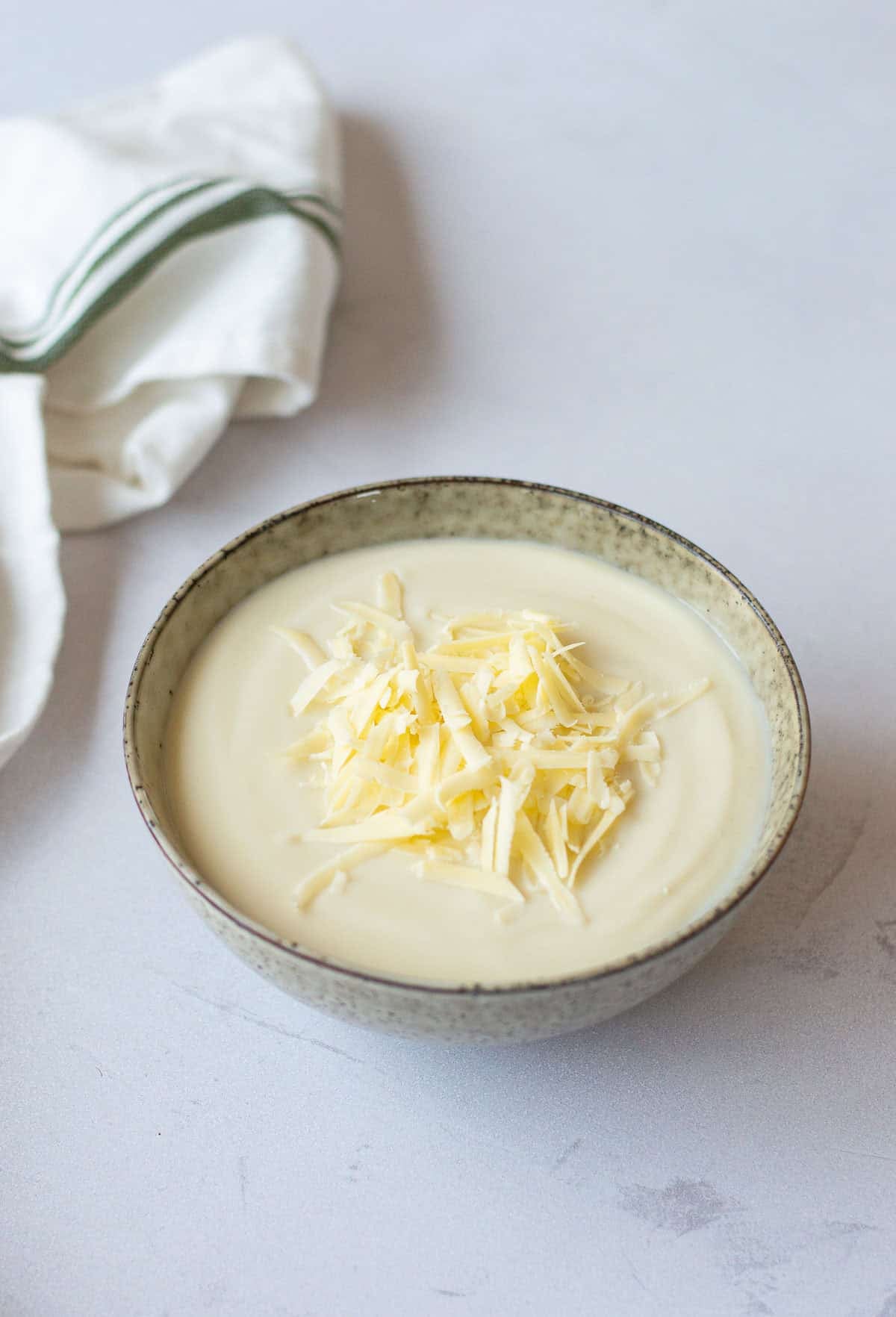A bowl of creamy cauliflower soup topped with grated cheese, placed on a light surface next to a white cloth with green stripes.