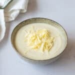 A bowl of creamy cauliflower soup topped with grated cheese sits on a light surface, with a white cloth serviette nearby.
