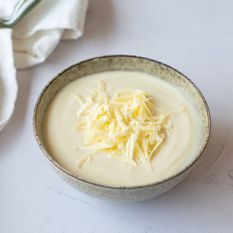 A bowl of creamy cauliflower soup topped with grated cheese sits on a light surface, with a white cloth serviette nearby.