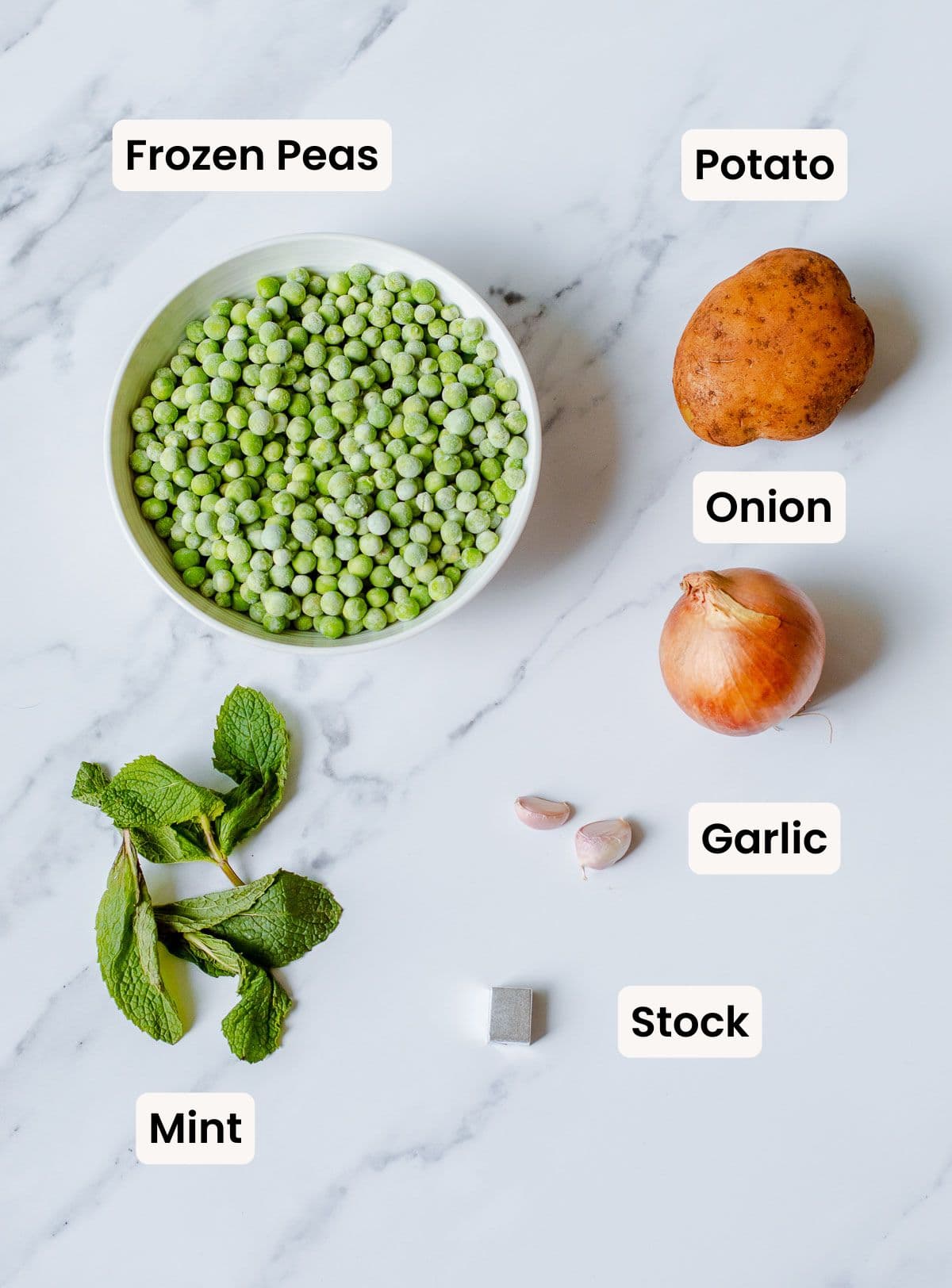 A bowl of frozen peas, a potato, an onion, two garlic cloves, a sprig of mint, and a stock cube are arranged on a white marble surface with labels identifying each ingredient.