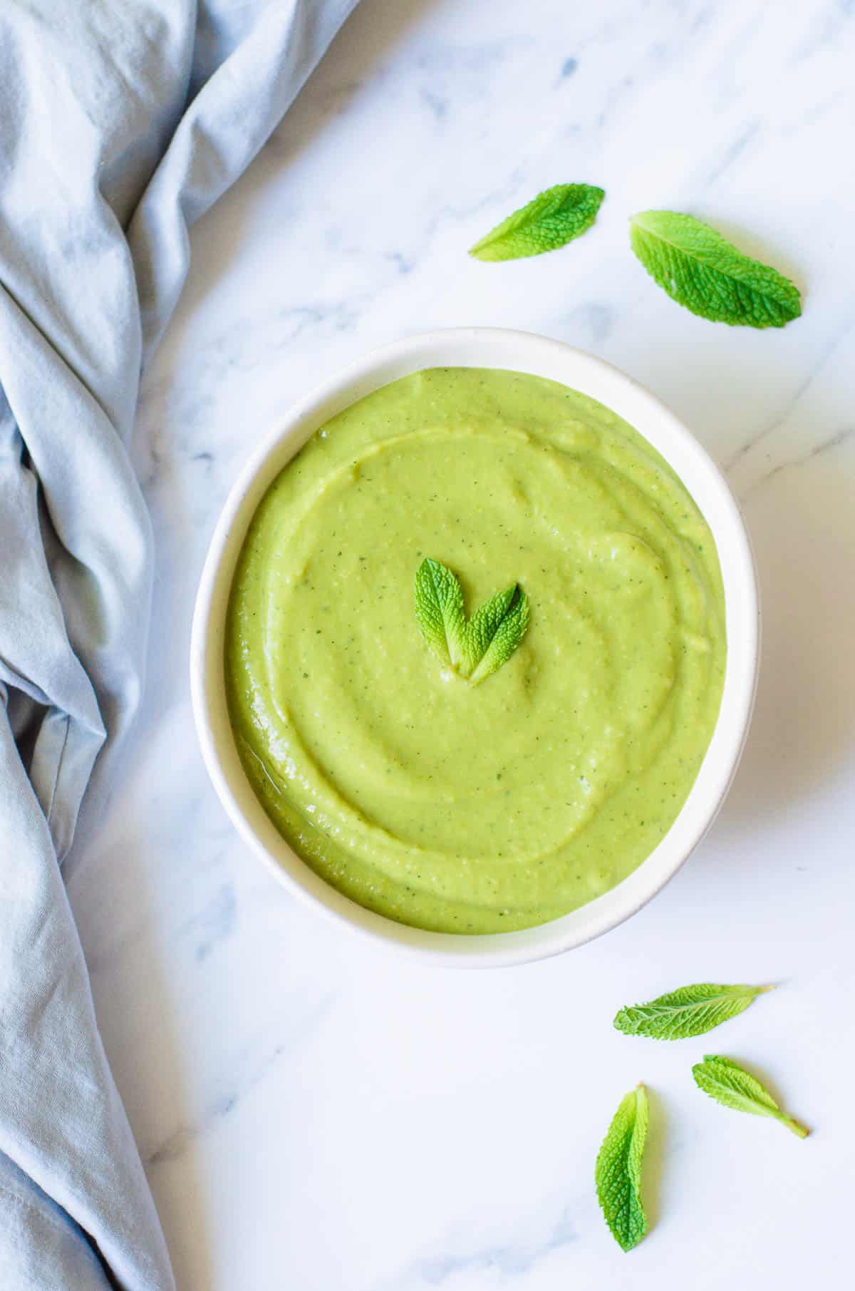 A white bowl filled with creamy pea and mint soup, garnished with two fresh mint leaves, sits on a marble surface with a few loose mint leaves and a light blue cloth nearby.