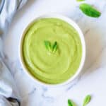 A white bowl filled pea and mint soup, garnished with two fresh mint leaves, sits on a marble surface with a few loose mint leaves and a light blue cloth nearby.