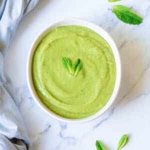 A white bowl filled pea and mint soup, garnished with two fresh mint leaves, sits on a marble surface with a few loose mint leaves and a light blue cloth nearby.